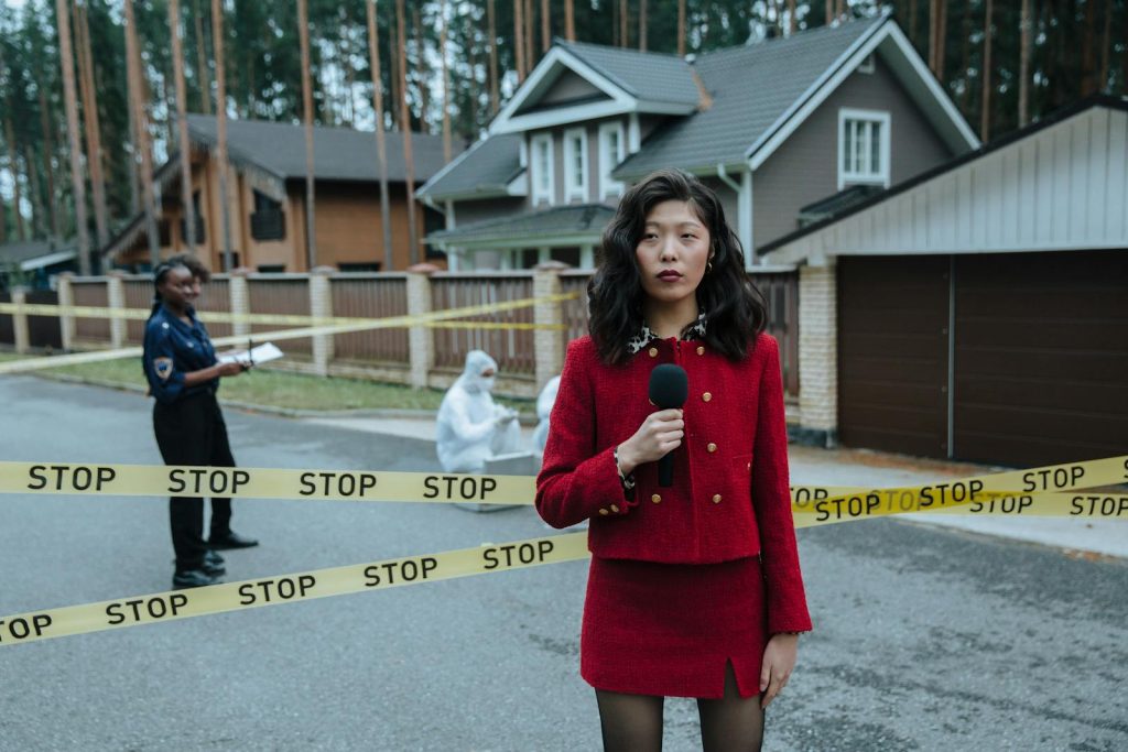 6 Aspects Worth Considering When Pitching Topics for Mobile Journalism, with 3 Examples 6 A journalist in a red outfit reporting live from a crime scene outdoors with stop tape and police presence.
