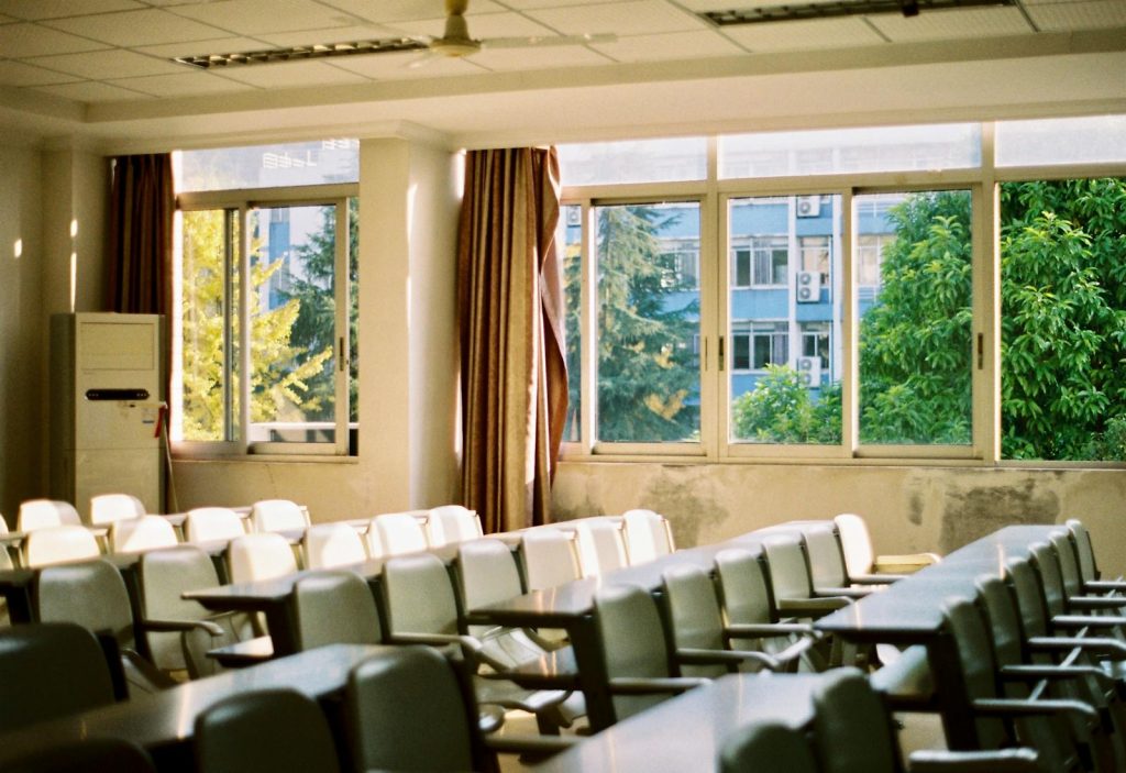 Bright empty classroom with rows of desks, chairs, and large windows overlooking greenery.