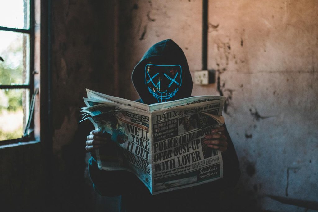 9 Rules to Identify Fake News on Online Platforms to Ensure You're Consuming Credible Content 5 A person in a neon mask and hoodie reads a newspaper in an eerie, abandoned room.