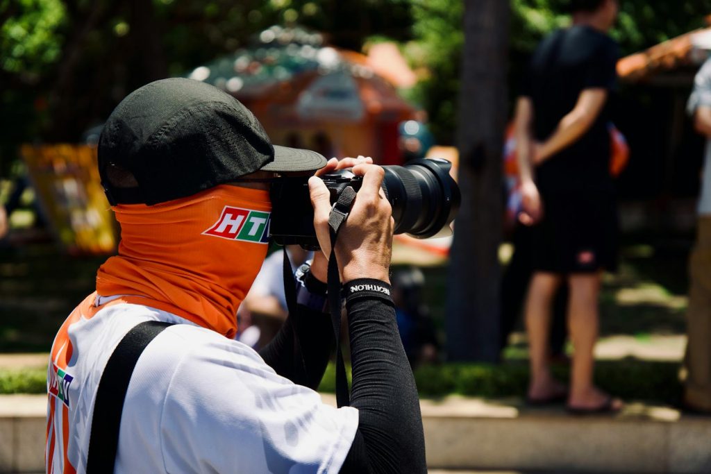 Masked photographer using DSLR camera at an outdoor event, focusing on capturing moments.