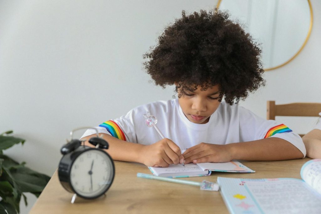 The Epitome Approach: Manage Academics and Extracurricular Activities Like a Charm 7 Concentrated African American child writing in notebook while studying at desk with alarm clock at home