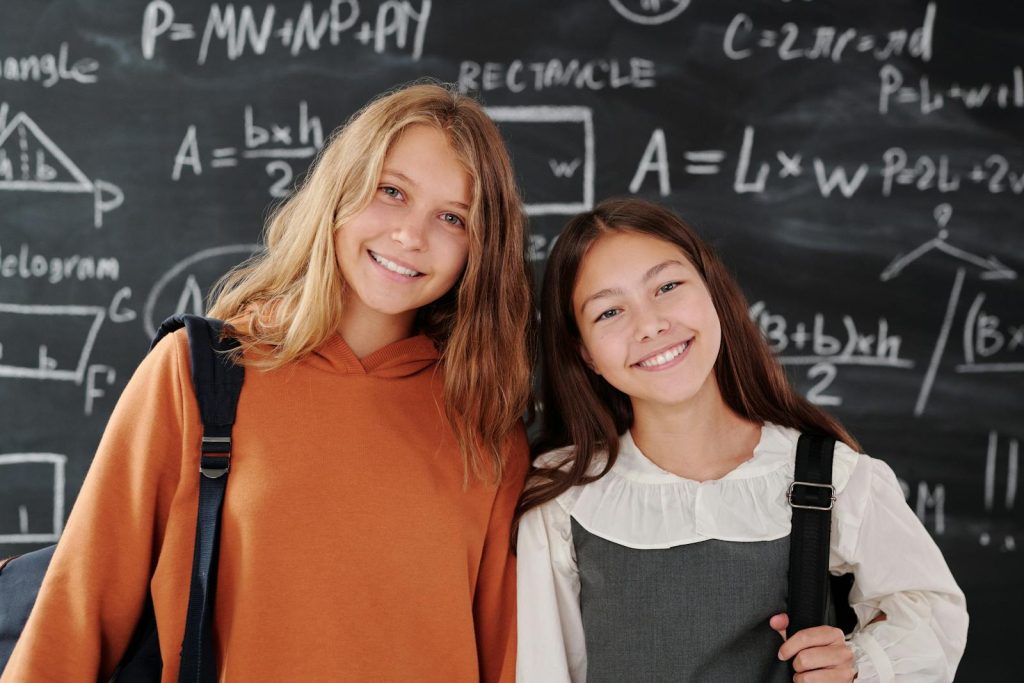 10 Simple Things to Soar Results with Classroom Management for Middle School Learners in the Digital Era 6 Two cheerful girls stand in a classroom with a math-covered chalkboard, ready for learning.