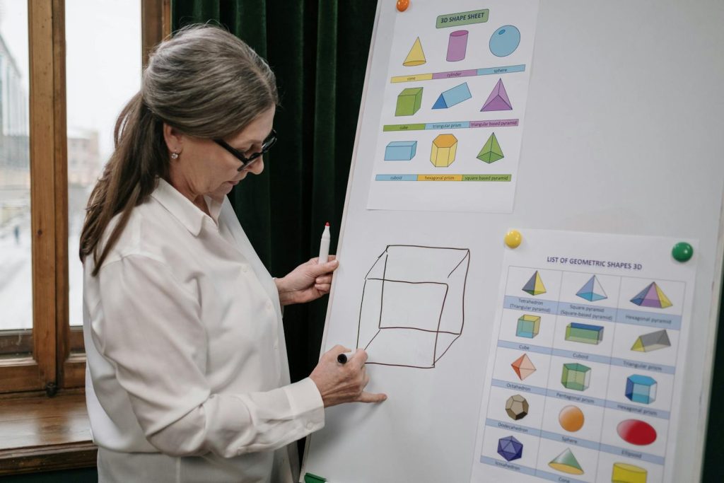 DepEd Partners with DOST for STEM Revolution, Scientific Innovations Underway 7 Senior woman instructing a geometry lesson using a whiteboard with 3D shapes.