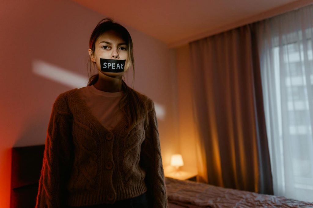 Rise of Teenage Pregnancy is Alarming, DepEd Commits to Health, Well-being of Youth 5 A woman stands in a dimly lit bedroom with tape reading 'Speak' over her mouth, symbolizing silence and oppression.