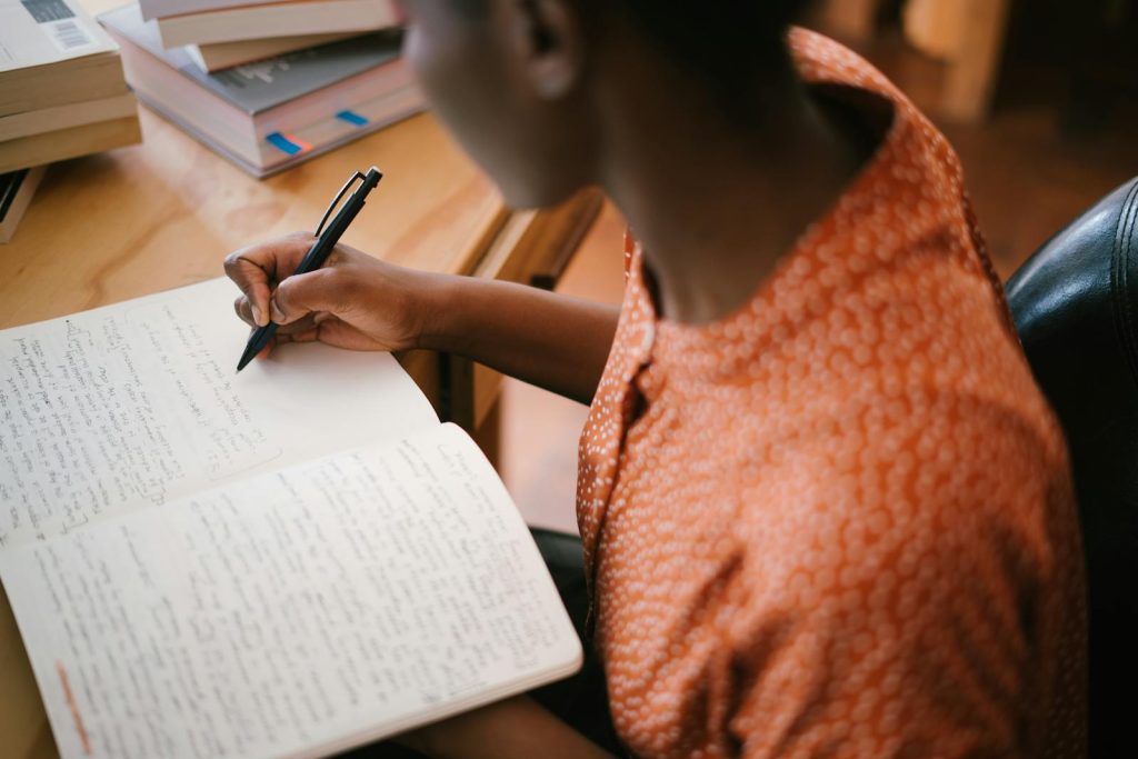 7 Extremely Fascinating New Year Study Habits Resolution for Kickass Results 5 Focused black woman writing in a notebook with stacked books on a desk, illustrating learning.