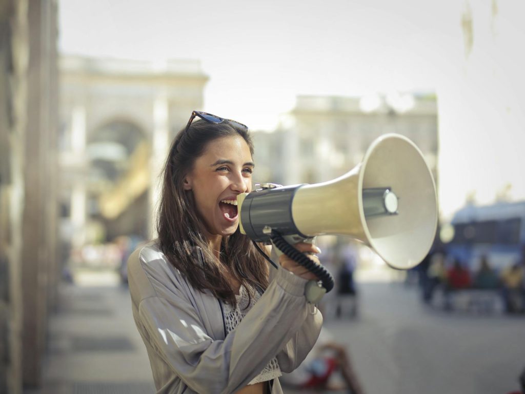 7 Empowering Editorial Writing Strategy for NSPC Victory 5 Cheerful young woman in a casual outfit shouting into a megaphone on a sunny day.