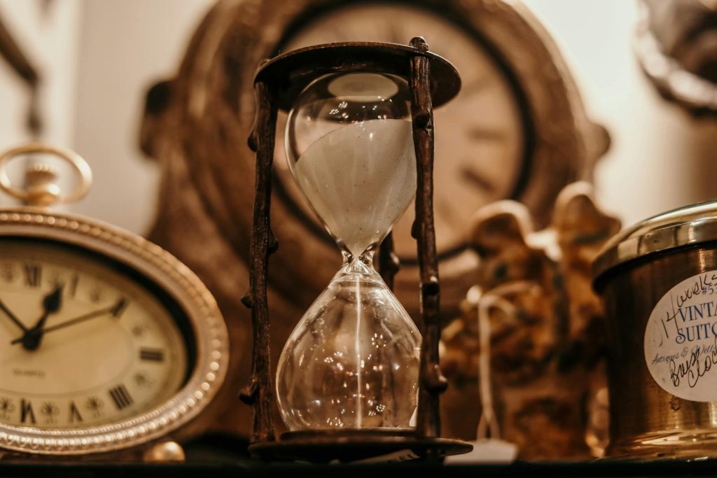 5 Easy Steps to Using the Pomodoro Technique, Game-Changing Tool For Procrastinators 8 Close-up of a vintage hourglass surrounded by antique clocks in warm lighting.