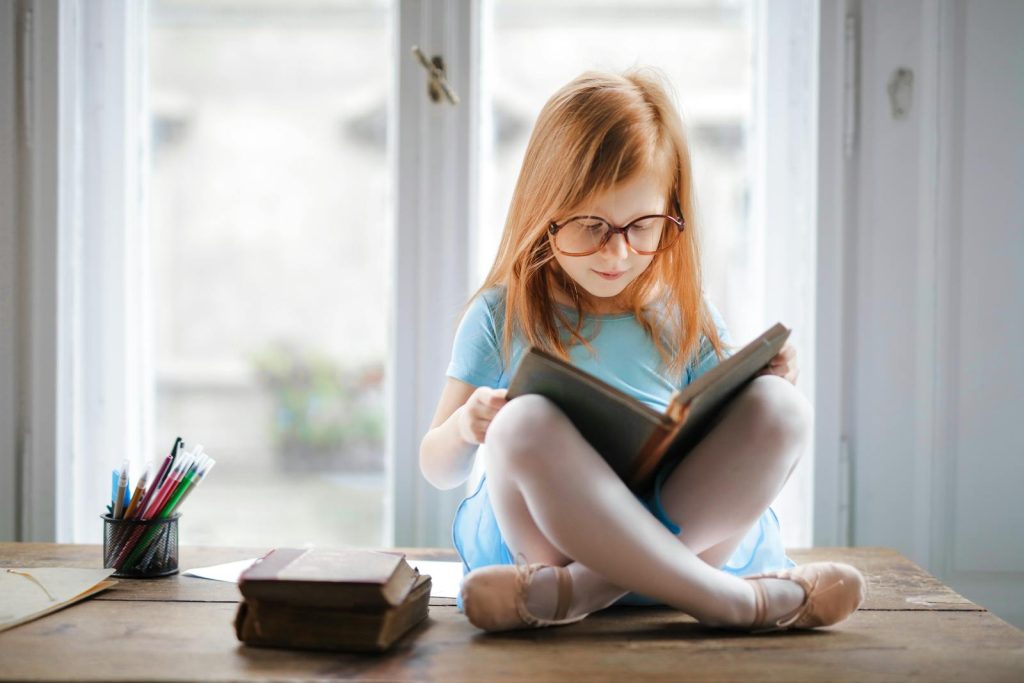 Interleaved Practice Makes You Learn Several Skills at Once 8 Adorable young girl wearing glasses, reading a book indoors by a window. Captures innocence and focus.