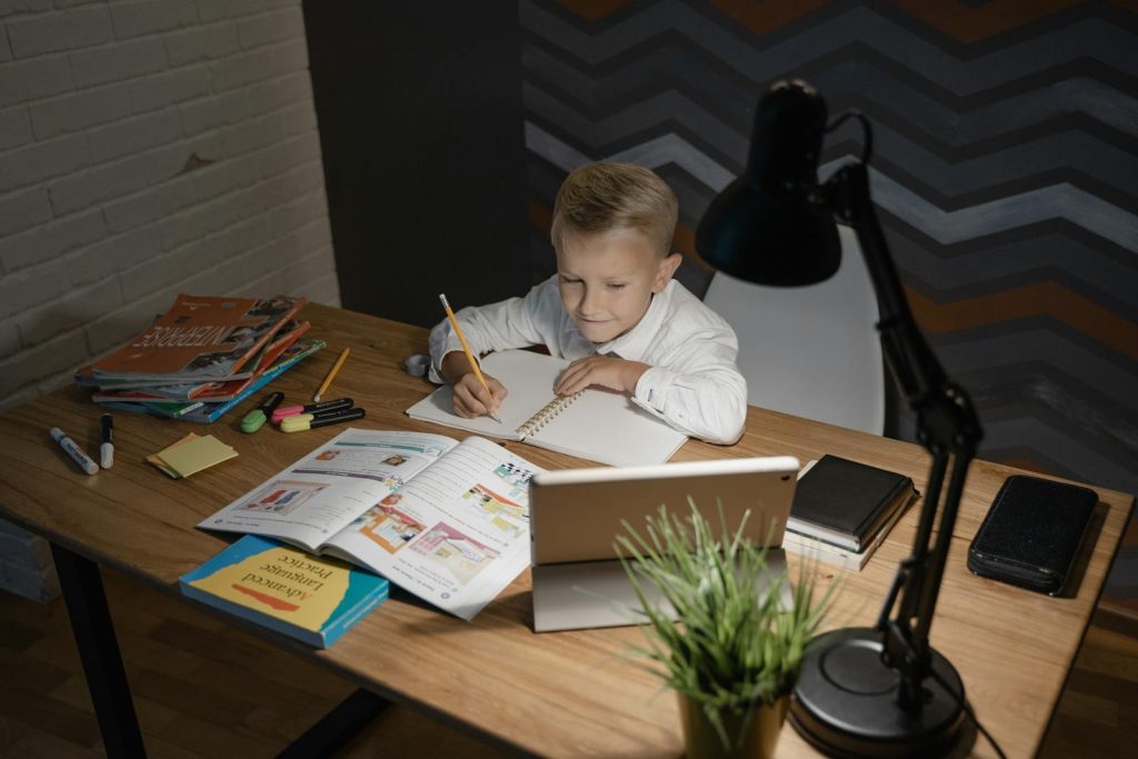 Interleaved Practice Makes You Learn Several Skills at Once 7 A young boy focused on studying at a wooden table with a tablet and books nearby under a desk lamp.