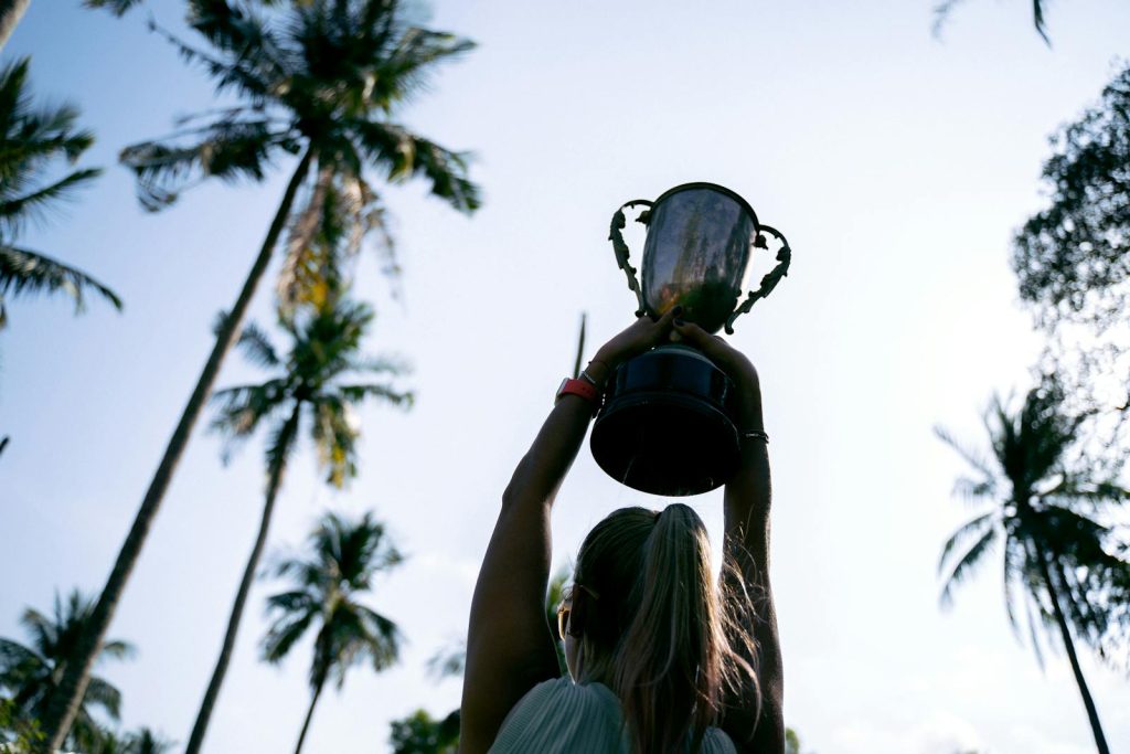 A woman lifts a trophy under palm trees, celebrating victory in nature.